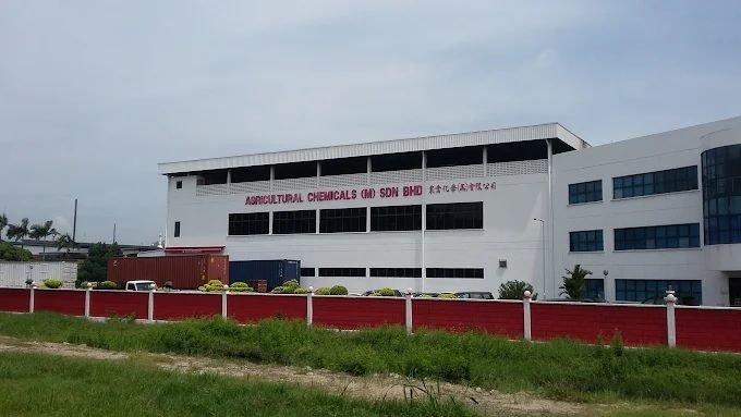 Exterior view of the white multi-story Agricultural Chemicals (M) Sdn. Bhd. manufacturing facility with red perimeter fencing, parked shipping containers, and company signage.