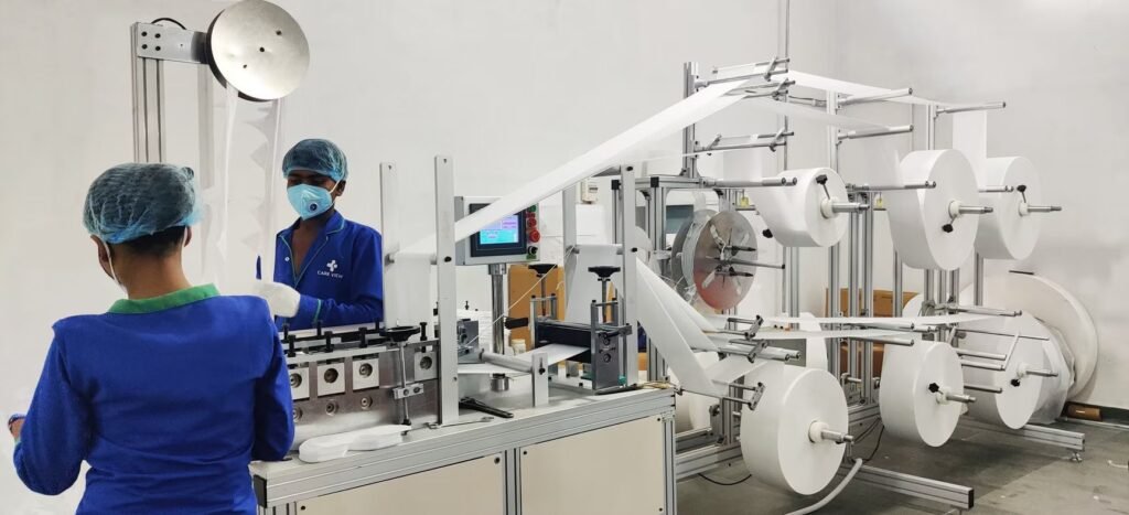 Factory workers wearing hairnets and protective face masks operating automated non-woven material production and converting machinery inside a clean industrial manufacturing facility.