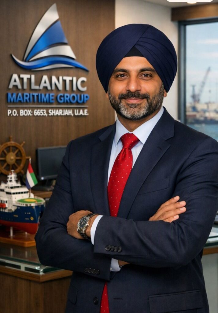 Corporate executive portrait of a Sikh professional wearing a dark turban, formal suit, and red patterned tie with arms crossed, set against the Atlantic Maritime Group wall logo, a ship model, and a port harbor window view.