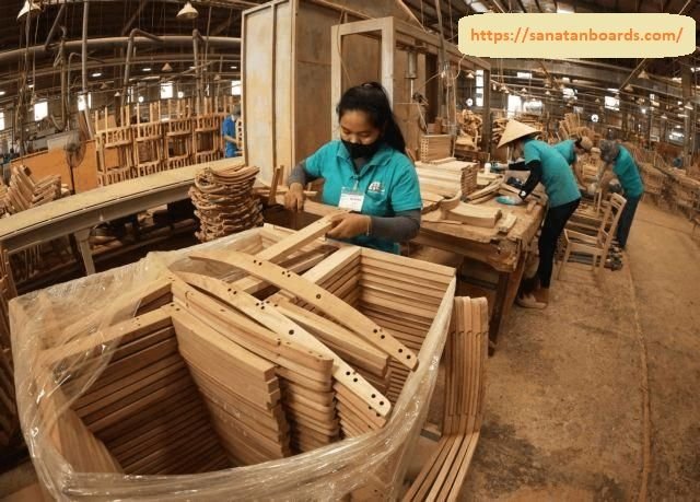 A factory worker in a teal shirt carefully assembling wooden chair components inside a busy furniture manufacturing facility.