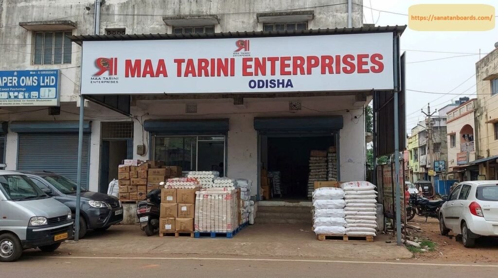 Exterior storefront view of Maa Tarini Enterprises in Odisha showing prominent red company signage, stacked cardboard boxes, paper roll bundles, sacks on pallets, and parked vehicles outside.