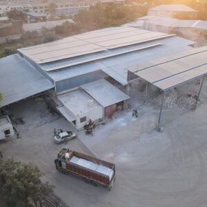 Aerial view of an industrial factory compound featuring metal-roofed sheds, loaded transport trucks, vehicles, and raw material storage areas.